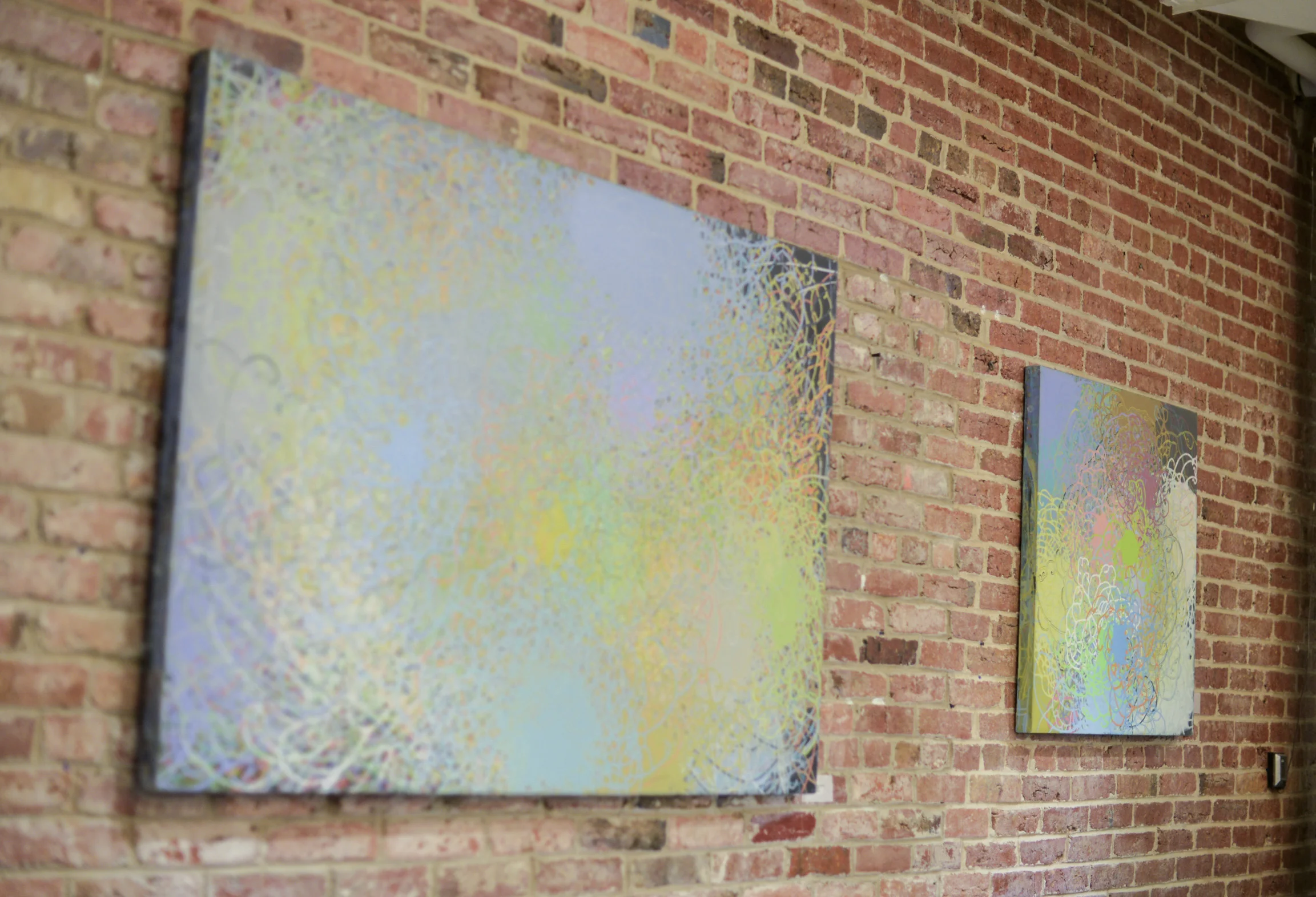 Two abstract paintings with pastel swirling patterns hung on an exposed brick wall, soft and airy mood