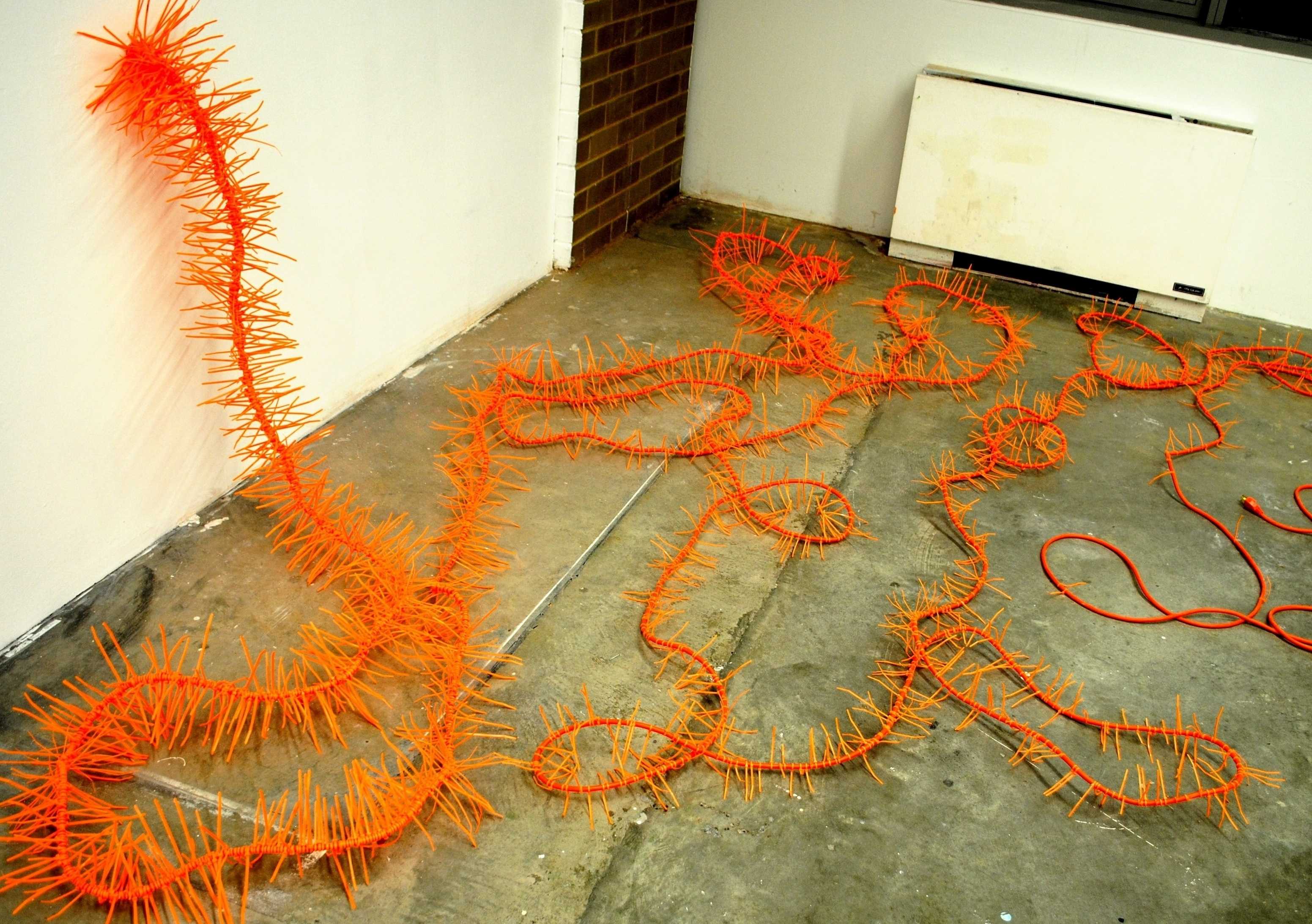 Orange spiky sculptural cord winding across a concrete floor and up a white wall in a bright, playful pattern